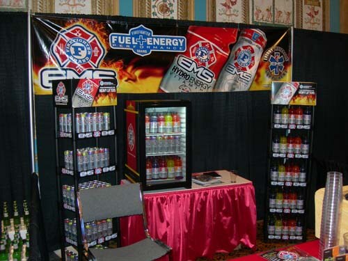 A booth with an EMS and Fuel+Energy banner above racks of canned and bottled drinks, a small lit refrigerator, and a table with a pink skirt. A gray chair is in front.