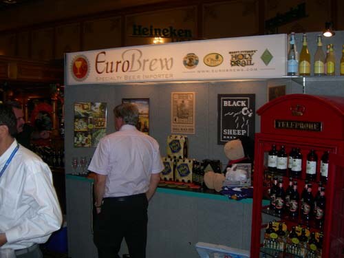 A booth with a EuroBrew sign, Holy Grail and Black Sheep posters, and a red telephone box displaying bottles. A man in a white shirt stands facing the booth.