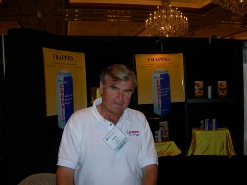 An older man in a white polo shirt and name badge stands in front of a black booth with yellow Frappio posters showing blue cans. A chandelier hangs overhead.