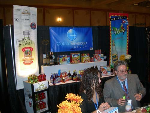 Gelosо Beverage Group booth with bottles and boxes on a draped table, a Popper's Mojito banner at right, and a seated man and woman wearing badges in the foreground.
