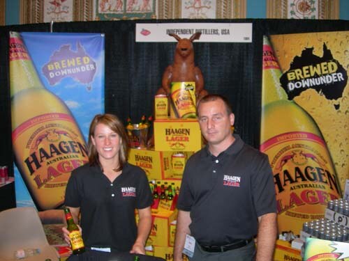 A woman holding a beer bottle and a man, both in black polo shirts, stand at the Independent Distillers, USA booth with Haagen Australian Lager banners and a kangaroo figure on stacked boxes.