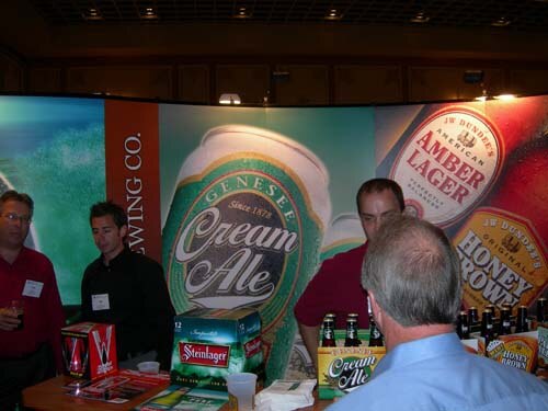 Booth with large Genesee Cream Ale and J.W. Dundee's banners, Steinlager and Cream Ale cartons on a table, and several men standing around, one seen from behind in a blue shirt.