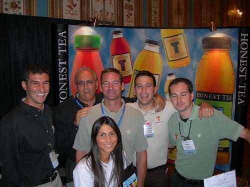 Five men and one woman pose close together in front of an Honest Tea backdrop showing large illustrated tea bottles, most wearing badges and smiling at the camera.