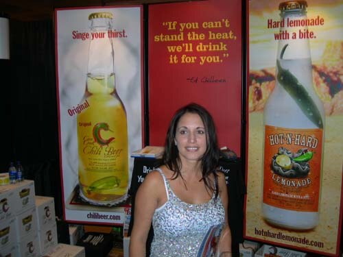 A smiling woman with long dark hair in a sparkly top stands between large posters for Chili Beer and Hot-N-Hard Lemonade, with boxes stacked at left.