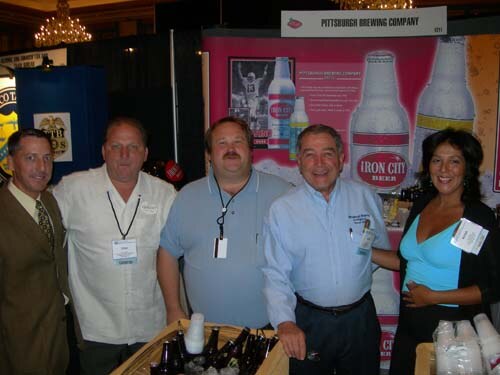 Four men and a woman pose together at the Pittsburgh Brewing Company booth in front of an Iron City Beer display, with bottles on ice in a wooden crate in front of them.