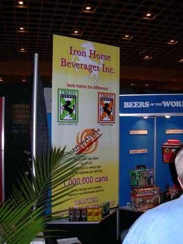 Tall yellow Iron Horse Beverages Inc. banner for Iron Energy with a horse logo and cans on a table, a palm plant in the foreground and a Beers of the World sign behind.
