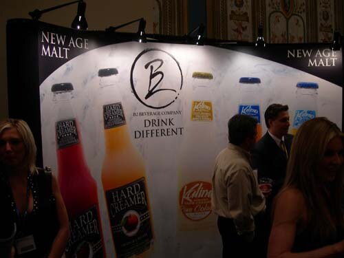 BJ Beverage Company backdrop reading Drink Different, with Hard Dreamer and Kalima bottle graphics, and several people standing in front of it in dim lighting.