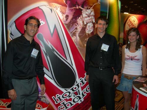 Two men in black shirts with name badges and a young woman in a white tank top stand in front of a red Liquid Charge banner with a lightning bolt design.