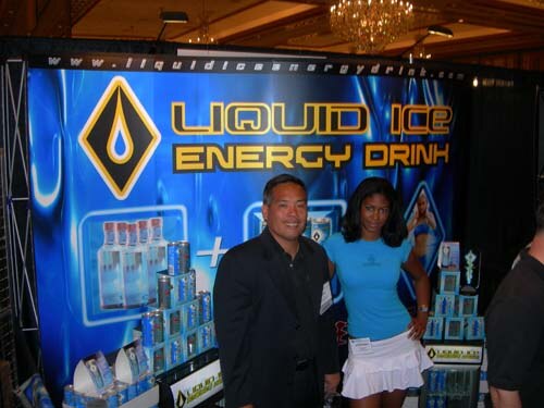 A man in a dark jacket and a woman in a light blue top and white skirt stand before a blue Liquid Ice Energy Drink backdrop with stacked cans and bottles.
