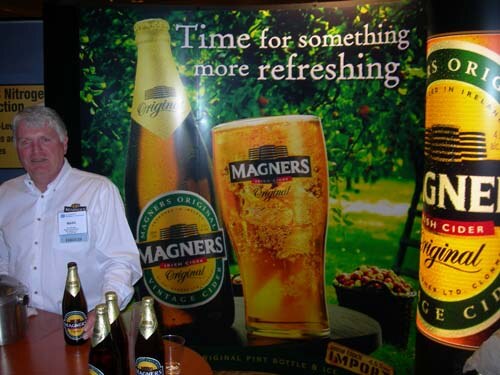 A gray-haired man in a white shirt and name badge stands behind a table with Magners cider bottles, in front of a banner reading 'Time for something more refreshing'.