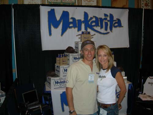 A smiling man in a cap and a blonde woman in a white Markarita shirt stand before stacked Markarita boxes and a blue Markarita banner.
