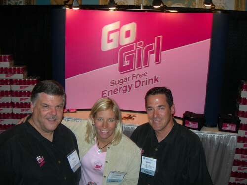 Two men in black shirts and a blonde woman in a pink top, all with badges, stand in front of a pink Go Girl Sugar Free Energy Drink sign with stacked cases.
