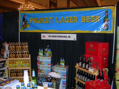 A booth with a yellow-bordered Phuket Lager Beer banner above a Best Brands International sign, with beer bottles and cases displayed on shelves and tables.