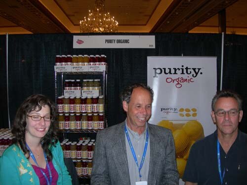 A woman with glasses and two men, all wearing lanyards, stand in front of shelves of bottled drinks and a Purity Organic banner with lemons.