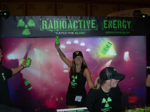 A woman in a black cap holds up a green can under a Radioactive Energy banner, with another woman in a cap in the foreground and a person holding up a can at left.