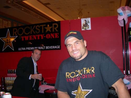 A smiling man in a Rockstar Energy Drink cap and shirt sits in front of a red wall with a Rockstar Twenty-One sign; a man in a dark suit stands behind him.