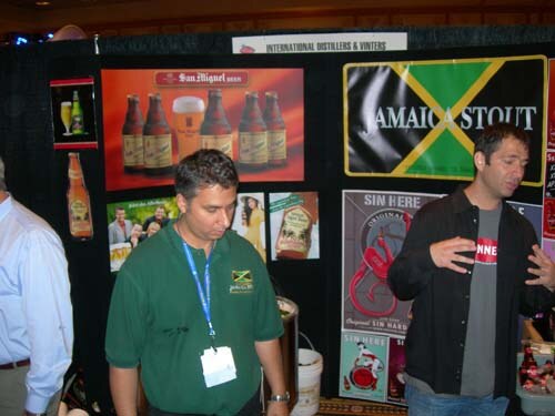 Two men stand at a black-draped booth with San Miguel beer posters and a green, yellow and black Jamaica Stout banner. A sign reads International Distillers & Vintners.