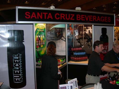 A Santa Cruz Beverages booth with a large FuelCell bottle graphic on the left, bottle displays on shelves, and a woman and two men standing at the counter.