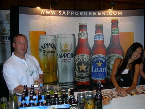 A man in a white shirt and a smiling woman stand behind a counter in front of a Sapporo backdrop showing Sapporo Reserve cans, Sapporo Light and other bottles, with beer bottles and cans on the table.
