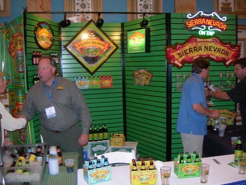 Men stand at a green-paneled Sierra Nevada booth with signs, including a lit Sierra Nevada On Tap sign, and six-packs of beer on the counter.