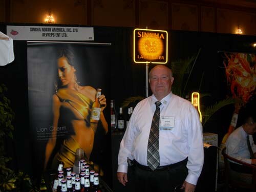 A man in a white shirt and striped tie stands in front of a Singha Lager Beer lit sign and a poster of a woman holding a bottle. A sign names Singha North America and Devreps Ent Ltd.