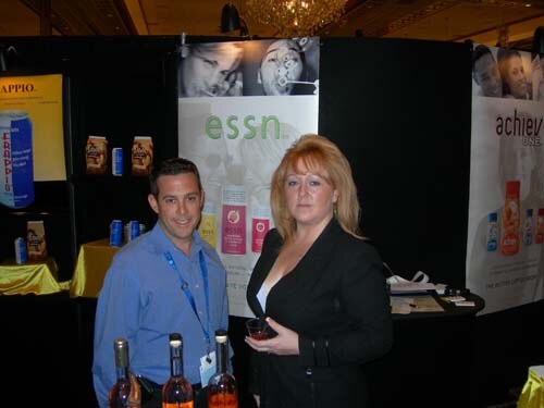 A man in a blue shirt and a woman in a black jacket holding a small glass stand at a booth with an ESSN banner. Bottles of orange-colored drink sit on the table in front of them.