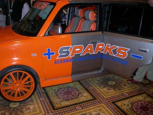 An orange and gray car with orange wheels on a patterned carpet, with the Sparks name and the words Alcohol/Caffeine/Cruise Hybrid on its side.