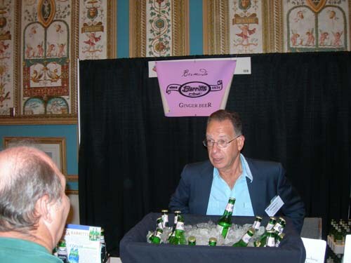A man in a blue blazer sits behind a tub of iced green bottles under a pink Barritt's Ginger Beer sign, while another man's head is in the foreground.