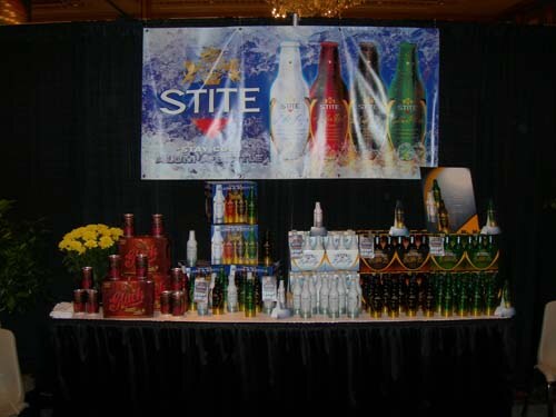 A black-draped table holds stacked cases and bottles of beer beneath a Stite banner showing four bottles, with yellow flowers at the left.