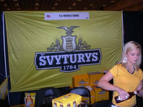 A woman in a yellow top pours a bottle of beer in front of a yellow Švyturys 1784 banner labeled B & I Overseas Trading, with cartons of beer stacked behind her.