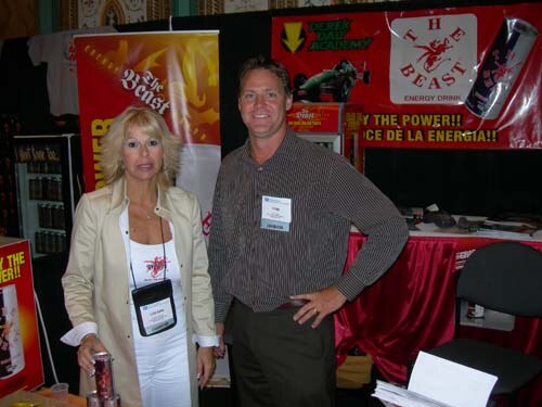 A woman in a cream jacket and a man in a striped shirt with a name badge stand together in The Beast energy drink booth, with Beast banners and a red tablecloth behind them.