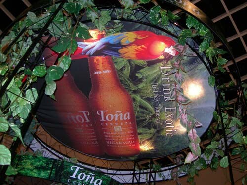 An overhead circular display shows a parrot and two Toña beer bottles with the text Drink Exotic, surrounded by green vines and a Toña sign below.