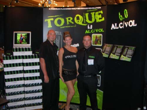 Two men in black and a woman in a black top and short skirt stand in the Torque booth, beside stacked cases of Torque cans and a banner reading 6.0 ALC/VOL.