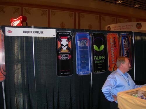 A man in a light blue shirt stands at the Unique Beverage, LLC booth, where tall can graphics for Alien Energy and Wired hang on a black curtain beneath a Miller sign.