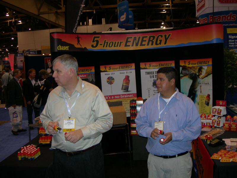 Two men with lanyards stand in the 5-hour ENERGY booth holding small bottles, with product posters behind them and bottles displayed on tables.