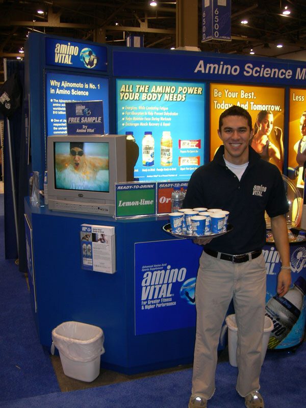 A smiling man in a dark Amino Vital polo holds a tray of sample cups in front of a blue Amino Vital booth with a television, product signs and ready-to-drink bottles.