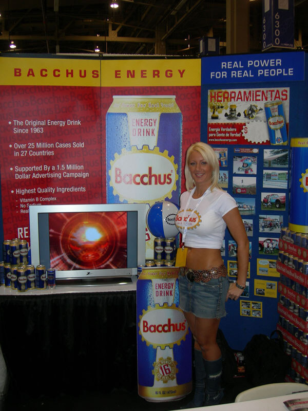 A blond woman in a white Bacchus top and denim skirt stands beside a large Bacchus energy drink can display, a television and a banner reading Bacchus Energy.