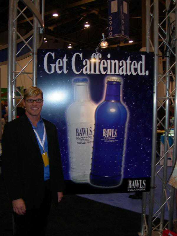 A man with glasses and a lanyard stands beside a large Bawls Guarana sign reading "Get Caffeinated." showing a clear sugar free bottle and a blue bottle, framed by metal truss.