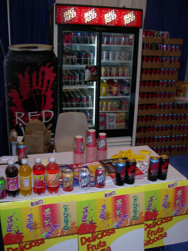 A booth table holds bottles and cans of Big Red, Red Jak and Nesbitt's drinks in front of a Big Red cooler, a giant Red Jak can and stacked cases of cans.