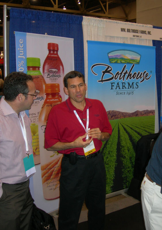 Two men talk in the Wm. Bolthouse Farms, Inc. booth, one in a red polo with an exhibitor badge, in front of Bolthouse Farms banners showing juice bottles and a carrot field.