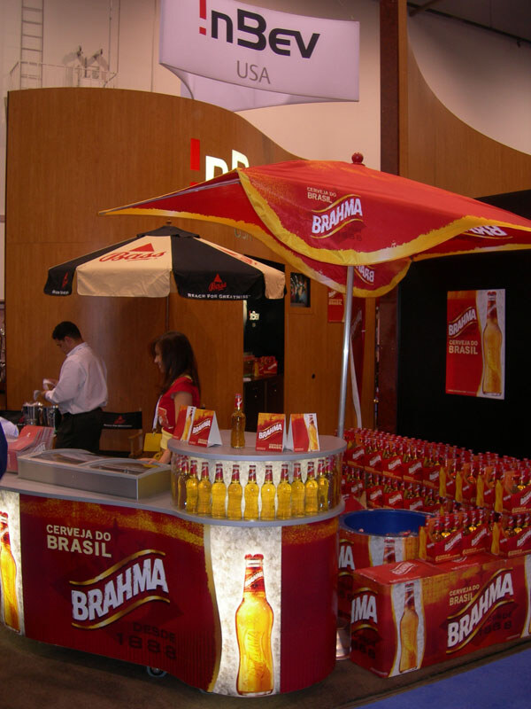 A Brahma beer booth under an InBev USA sign has a red Brahma umbrella, a lit counter with bottles, stacked Brahma cartons, and two staff members behind the counter.