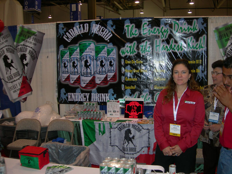 A woman in a red shirt with an exhibitor badge stands in the Caballo Negro Black Horse booth, with a black energy drink banner, cans on the table and two other people at right.