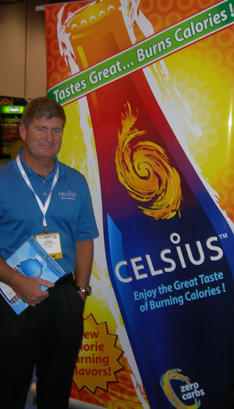 A man in a blue Celsius polo and lanyard holds a magazine while standing beside a tall Celsius banner reading "Tastes Great... Burns Calories!"