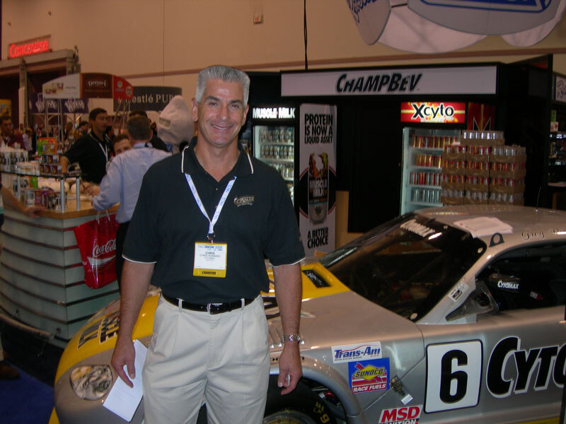 A smiling gray-haired man with an exhibitor badge stands in front of a ChampBev booth with Muscle Milk and Xcyto signs and a race car carrying Cytomax, Trans-Am, Sunoco and MSD decals.