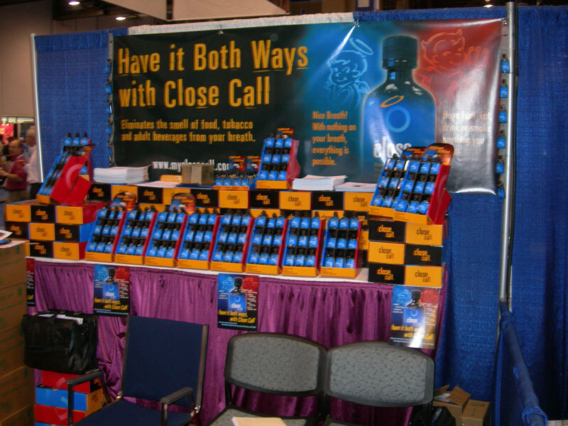 A booth with a blue backdrop and banner reading "Have it Both Ways with Close Call" shows many blue bottles in orange and black display boxes on a draped table.