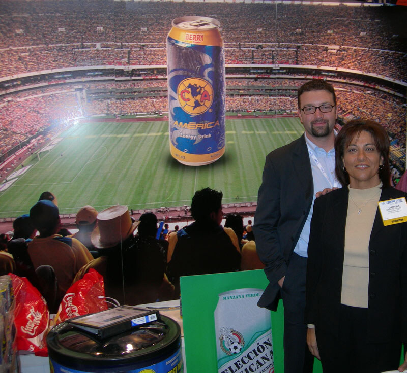 A bearded man with glasses and a woman with an exhibitor badge stand before a stadium backdrop with a giant Club América energy drink can.