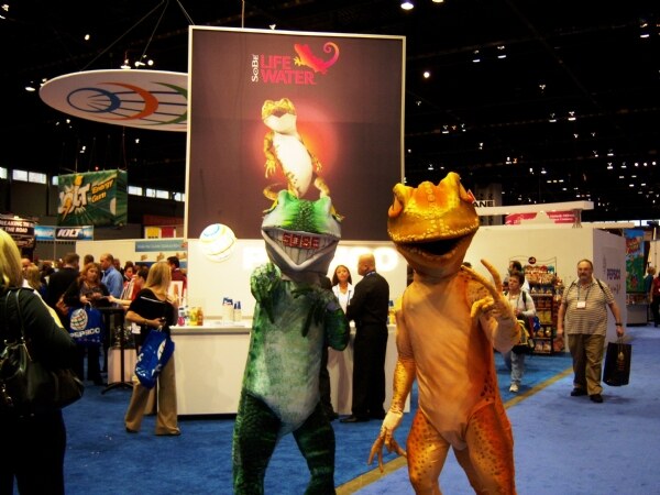Two people in lizard costumes, one green and one orange, stand on blue carpet in front of a SoBe Life Water booth sign. Attendees and other booths are visible behind them.