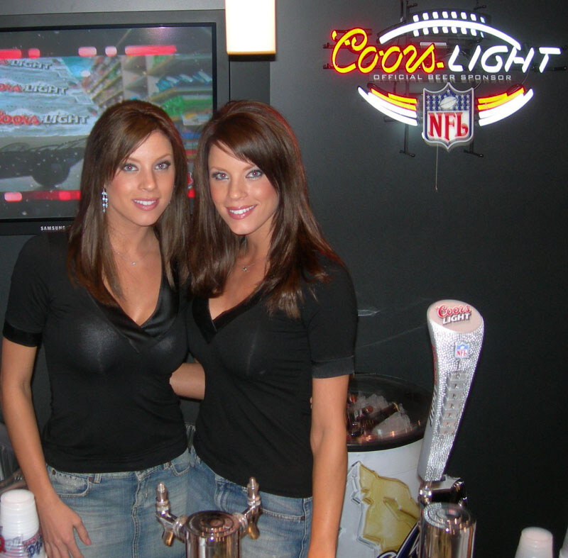 Two smiling women with long brown hair in black tops stand behind a bar with taps, in front of a lit Coors Light NFL neon sign and a Samsung screen showing Coors Light.
