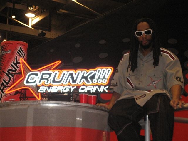 A man with long dreadlocks and white sunglasses, wearing a gray shirt, sits beside a lit Crunk!!! Energy Drink neon sign, a large can graphic and two red cups.