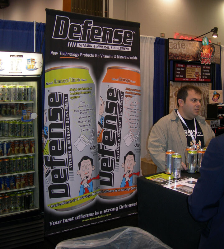 A man with a lanyard stands beside a tall Defense vitamin and mineral supplement banner showing Lemon Lime and Orange cans; a Planet Java coffee booth and a drink cooler are behind him.